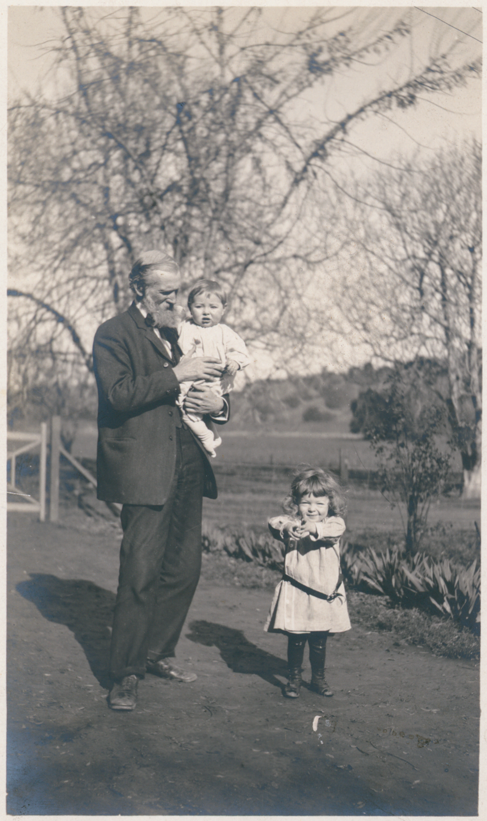 An elderly John Muir holding an infant grandchild while a young girl stands beside him in the ranch orchard.